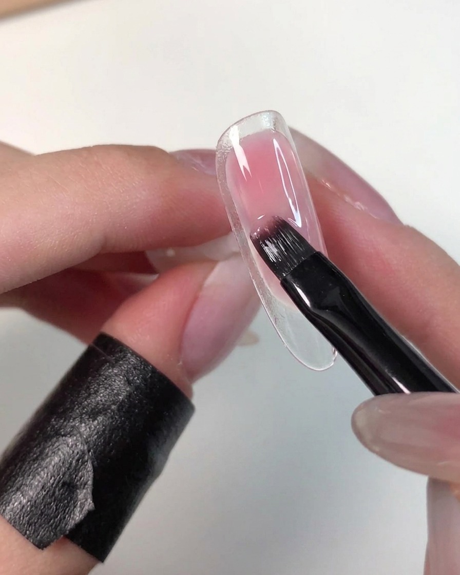 Eine Hand hält eine klare Nagelspitze, während ein flacher Pinsel feuchtes rosa Gel sanft einklopft, um die Ränder zu soften.