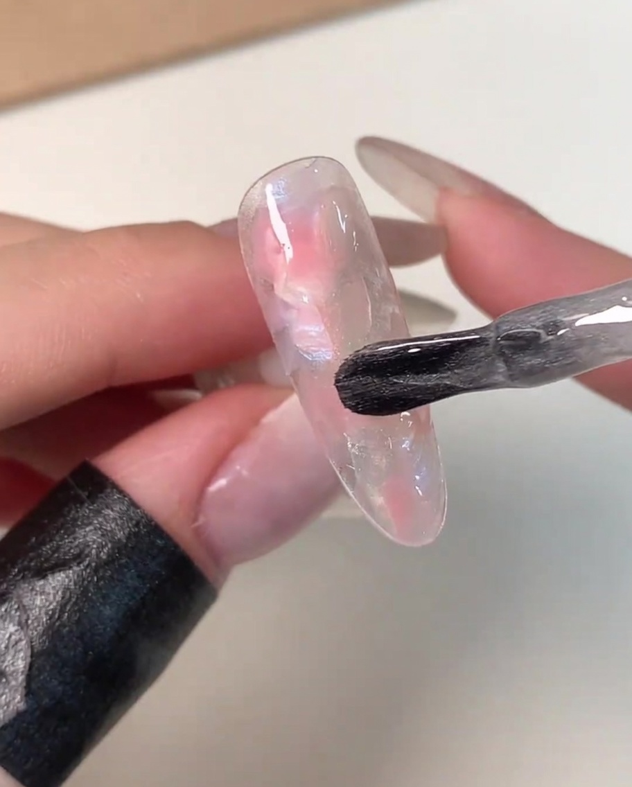 Ein Flaschenpinsel trägt eine Schicht klares, schimmerndes Magnetgel über einer rosa-weiß marmorierten Nagelspitze auf.