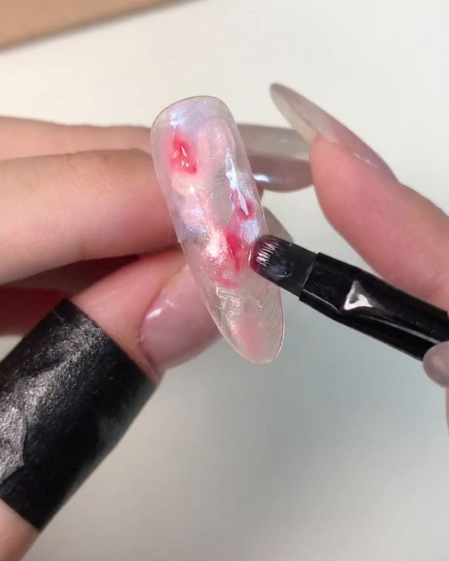 Ein kleiner flacher Pinsel tupft dunkleres rosa Gel in die Mitte einer nicht ausgehärteten, marmorierten Nagelspitze.