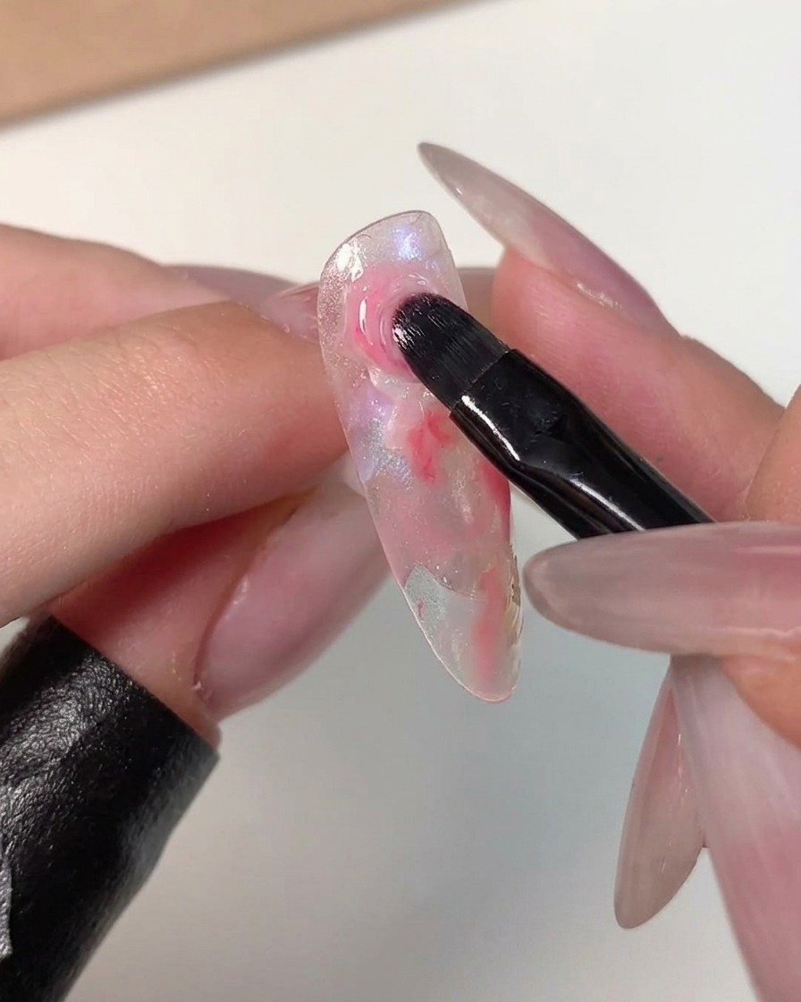 Ein Nail-Art-Pinsel federt die Ränder eines dunkelrosa Punkts in die umliegenden helleren Farben auf einer klaren Nagelspitze aus.