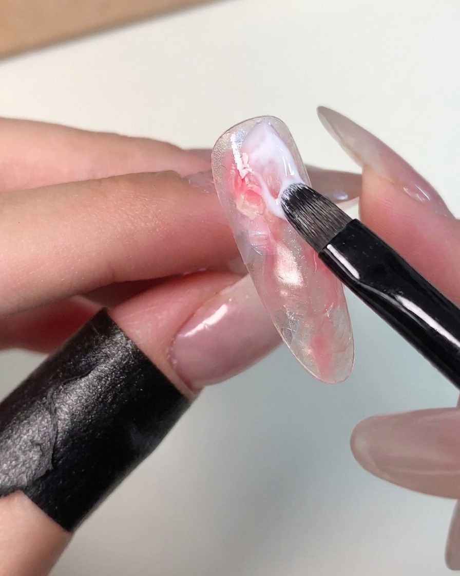 Ein kleiner Gelpinsel trägt einen Strich milchiges Weißgel nahe dem Nagelhautbereich einer marmorierten Nagelspitze auf, um das Muster zu verfeinern.