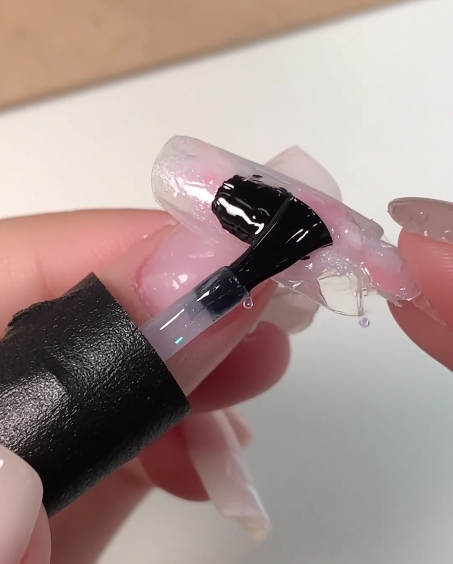 Ein schwarzer Nagelpinsel trägt eine dicke Schicht klares Leveling-Gel über ein marmoriertes rosa, weißes und blaues Nageldesign auf.