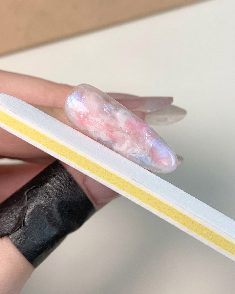 Eine Hand hält einen weiß-gelben Nagelfeilenstreifen und bufft die Oberfläche eines ausgehärteten, glänzenden marmorierten Gelnagels.