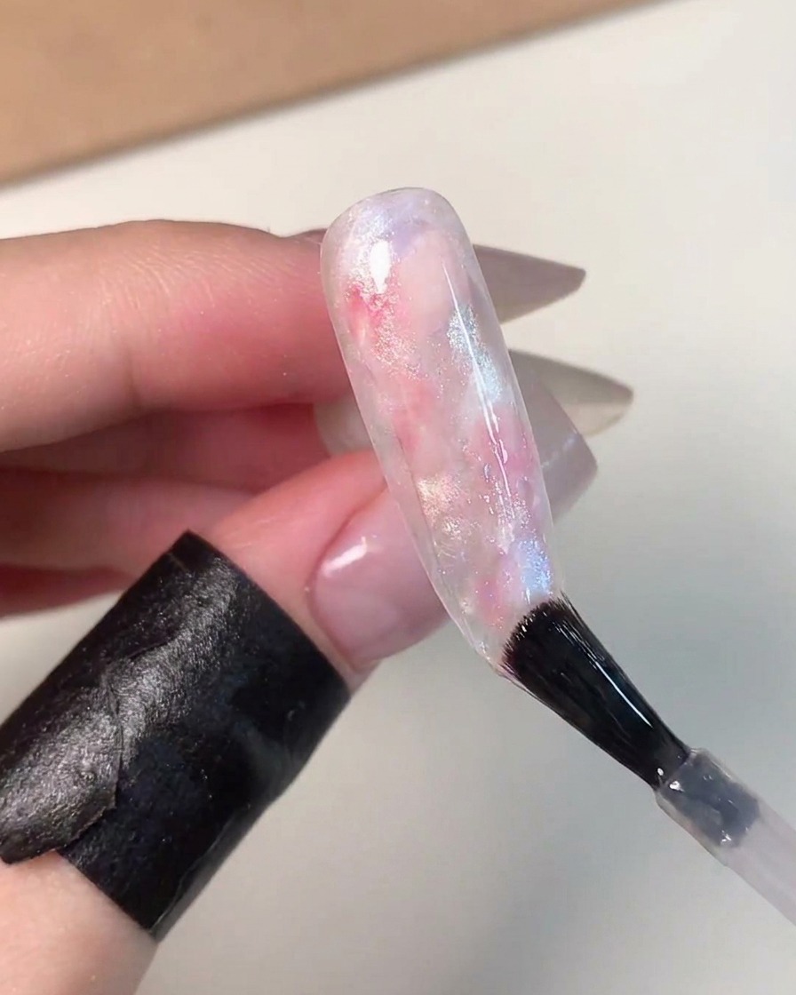 Ein Nagelpinsel trägt einen klaren, glänzenden Top Coat über einen wunderschön vollendeten rosa, blauen und weißen marmorierten Glaskugeleffekt-Nagel auf.