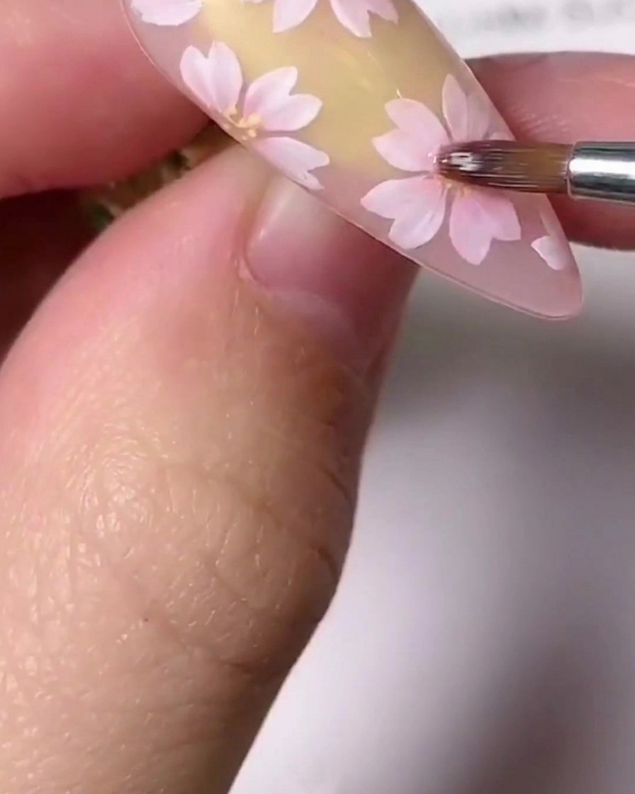 Eine Nahaufnahme eines feinen Detailpinsels, der kleine goldene Akzente in die Mitte einer rosa gemalten Kirschblüte auf einem Nagel setzt.
