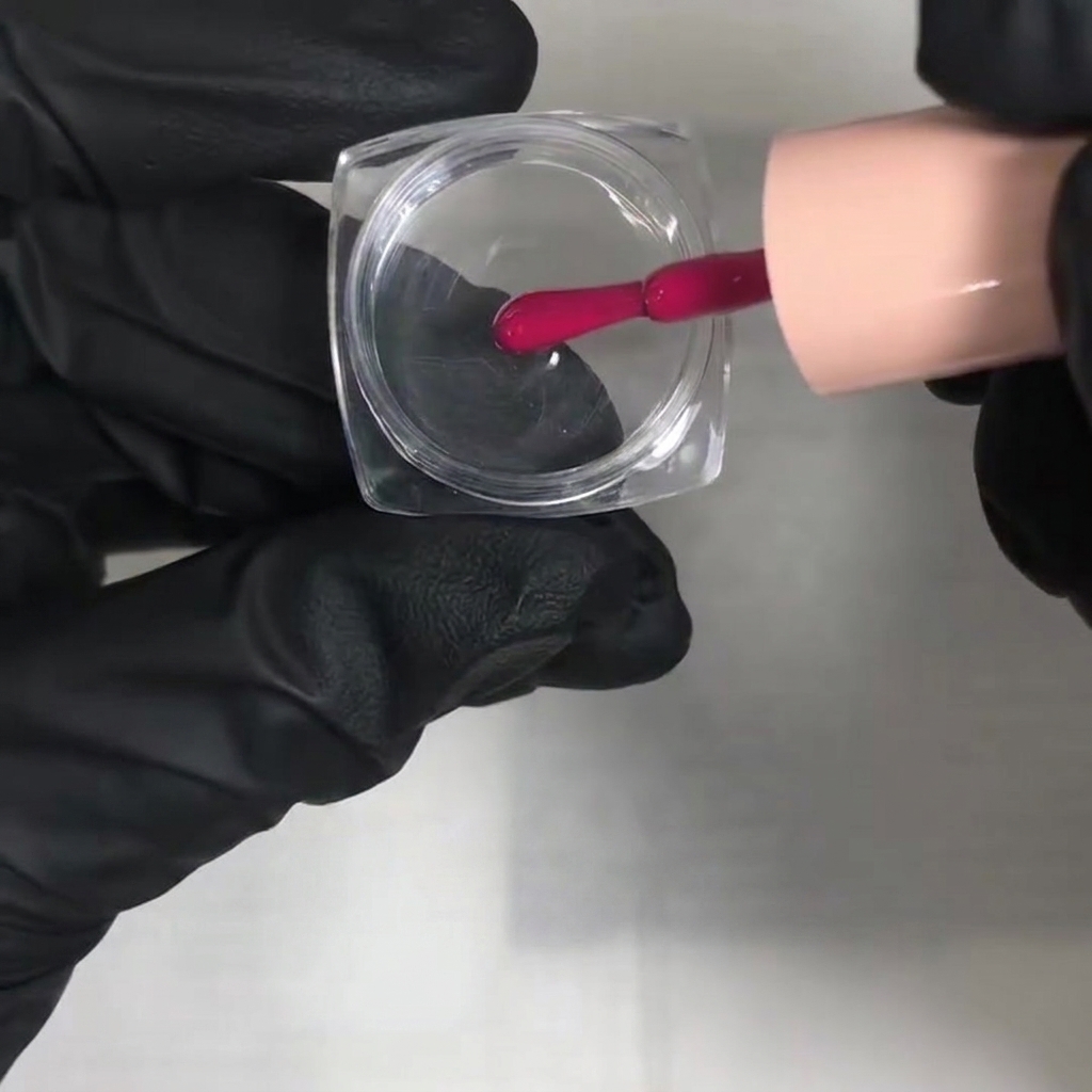Eine behandschuhte Hand gibt mit einem Pinsel leuchtend rosa Gel-Lack in ein durchsichtiges quadratisches Mischgefäß.
