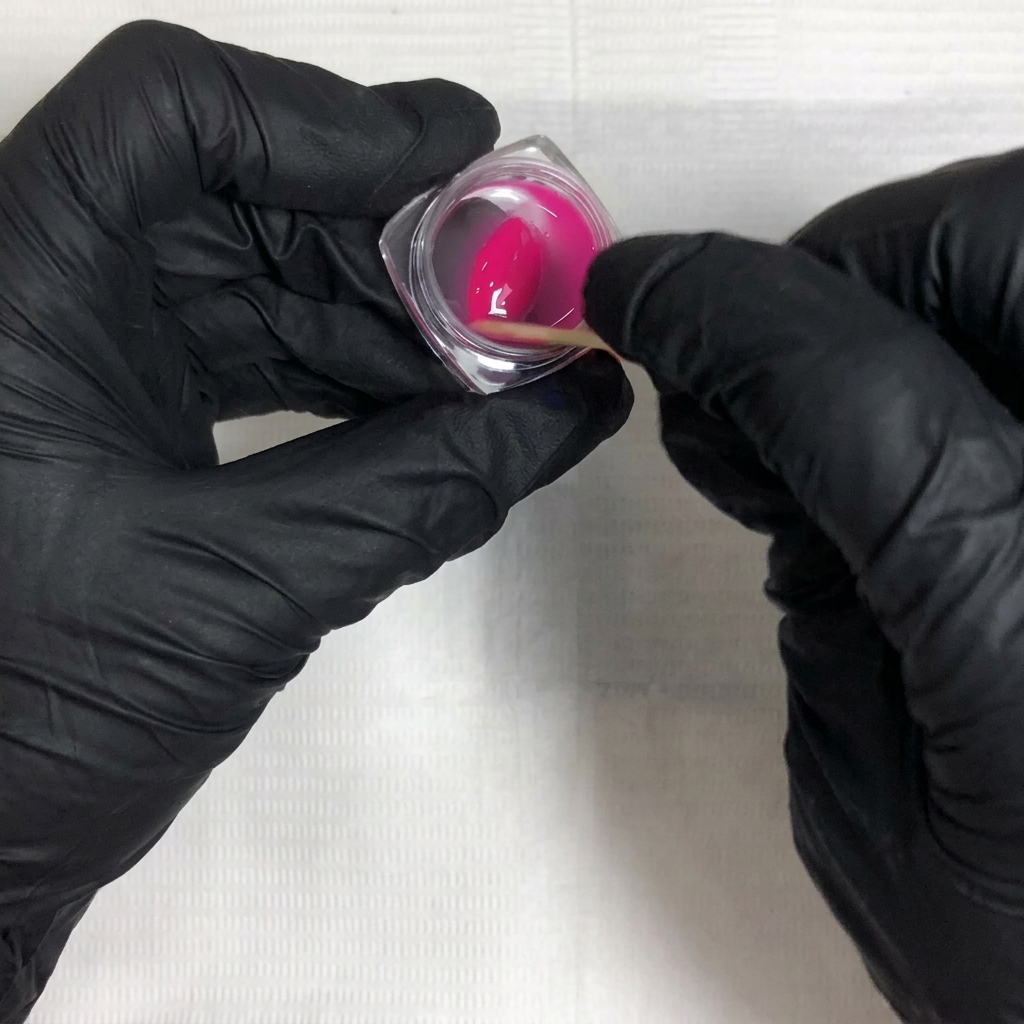 Zwei behandschuhte Hände mischen mit einem Holzstäbchen rosa Gel-Lack und flüssigen Verdünner in einem durchsichtigen Behälter.