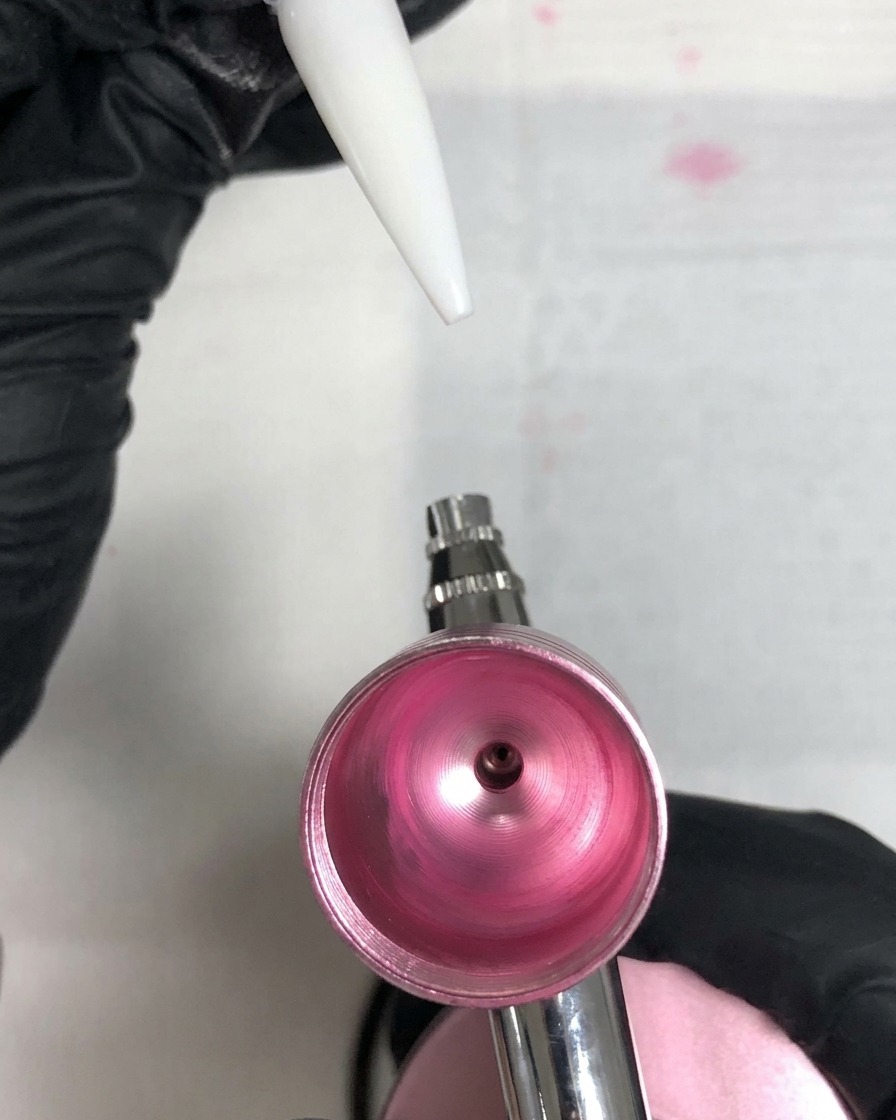 Ein weißer Nageltip wird über eine rosa Airbrush gehalten und zeigt einen dicken, ungleichmäßigen rosa Klecks durch falsches Sprühen.