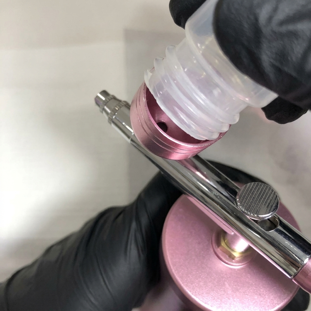 Behandschuhte Hände drücken eine klare Reinigungslösung aus einer Plastikflasche in den oberen Farbbecher einer Metall-Airbrush.