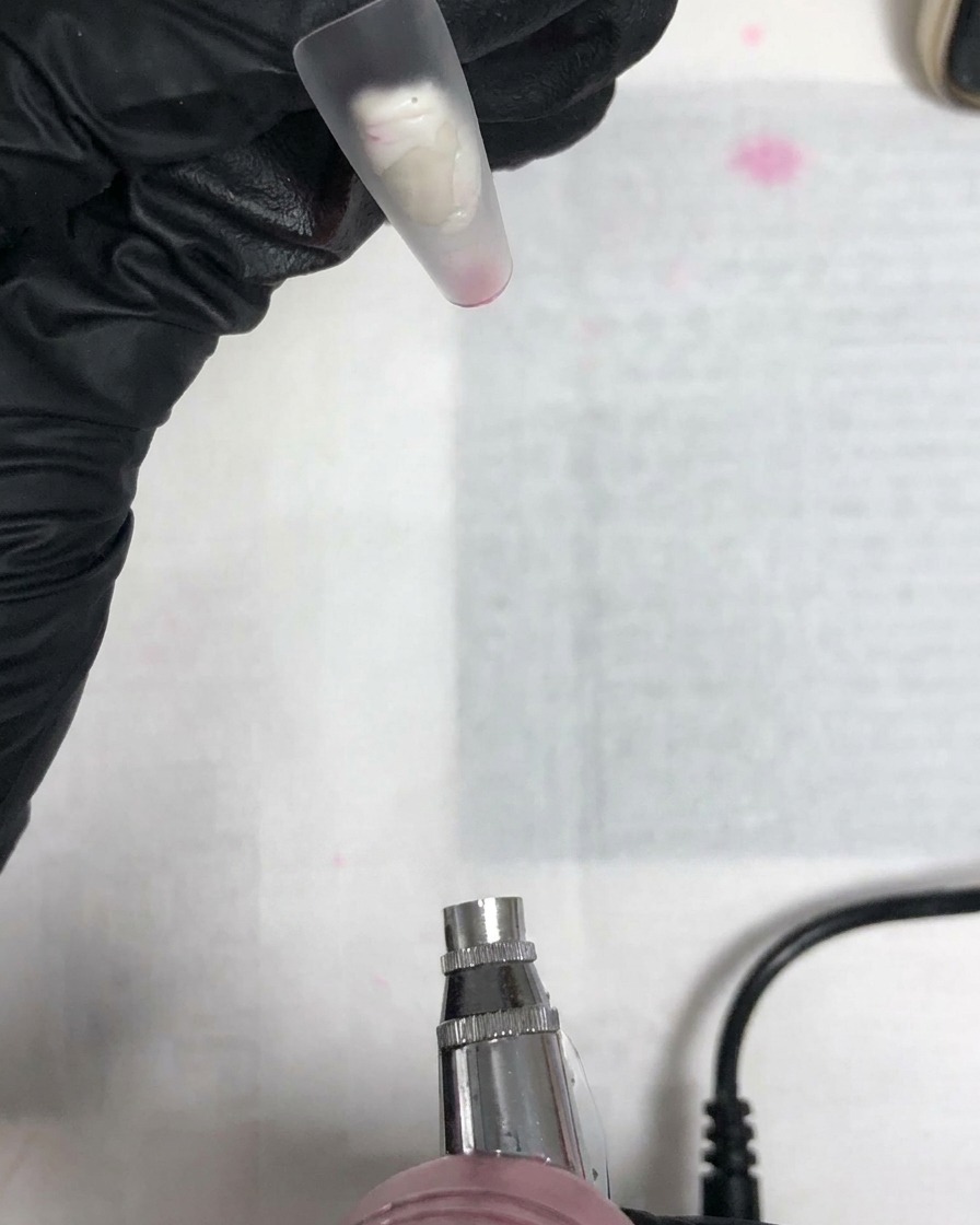 Eine Airbrush-Düse zeigt auf einen klaren Nageltip, der von einer behandschuhten Hand gehalten wird.