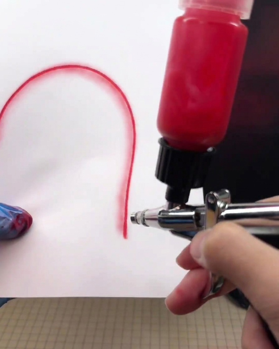 Eine Airbrushpistole, die sehr nah über ein weißes Papier gehalten wird und eine dicke, kräftige rote Linie zieht.