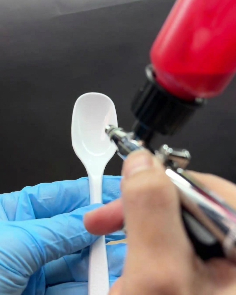 Eine Hand im Handschuh hält einen weißen Löffel, während eine rote Airbrush-Farbe aufgesprüht wird, wodurch ein auslaufender Farbverlauf entsteht.
