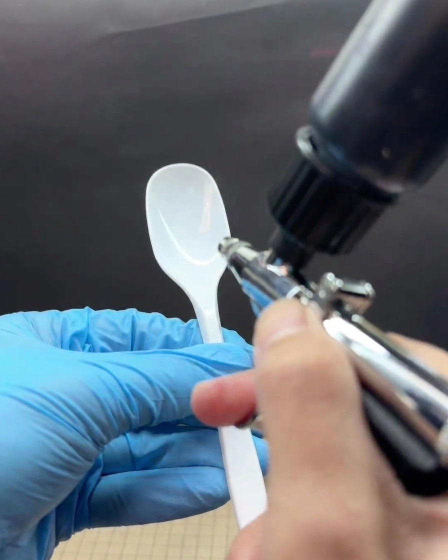 Eine behandschuhte Hand besprüht die Spitze eines weißen Löffels mit schwarzer Airbrush-Farbe und formt einen Farbverlauf.