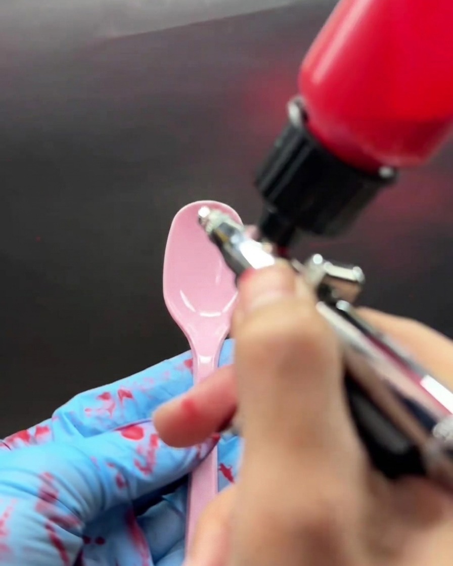 Eine behandschuhte Hand sprüht mit einer Airbrushpistole aus Metall eine pink-rote Farbe auf einen Plastiklöffel.