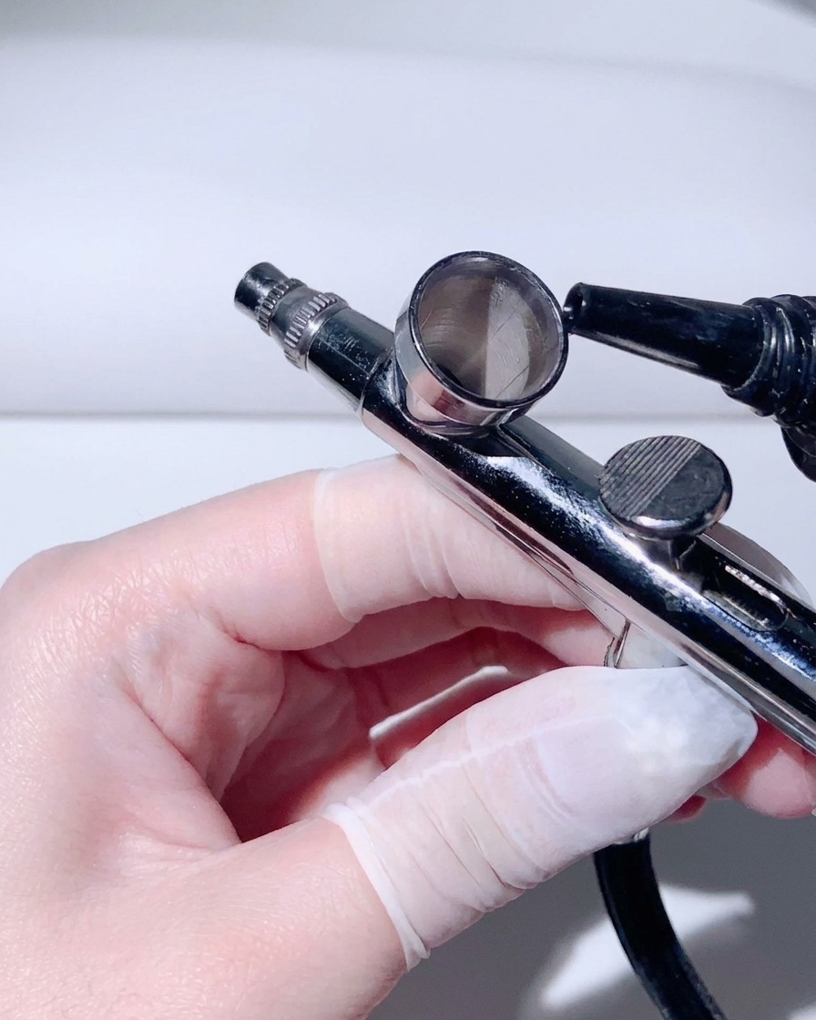 Eine behandschuhte Hand hält eine Chrom-Airbrush und gießt Verdünner in den kleinen Metallbecher.