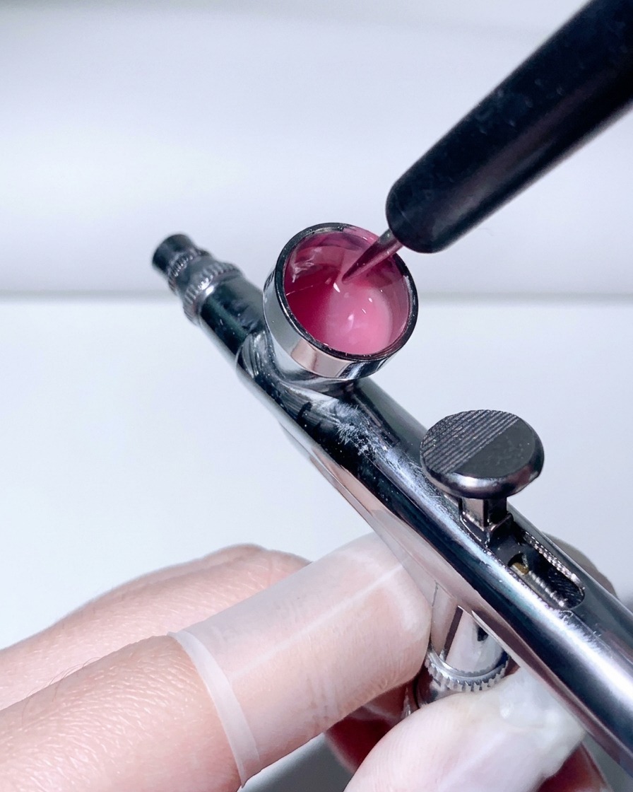 Ein Dotting-Pen mischt pinkfarbene Flüssigkeit im Metallbecher einer Airbrush.