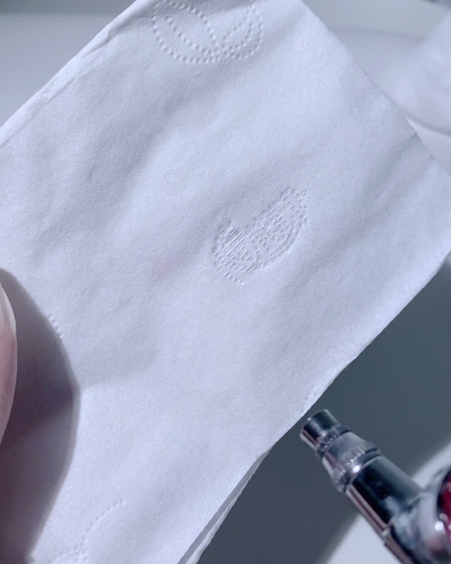 Eine Airbrush sprüht einen hellen, gleichmäßigen Pinknebel auf ein weißes Papiertaschentuch.