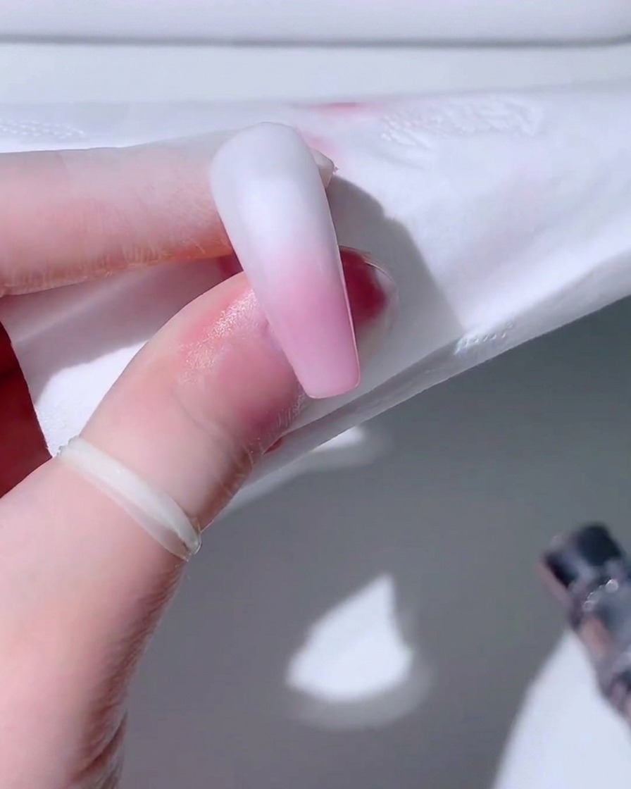 Ein Airbrush-Gerät nebelt sanft pinke Farbe auf den unteren Teil einer matten weißen Nagelspitze.