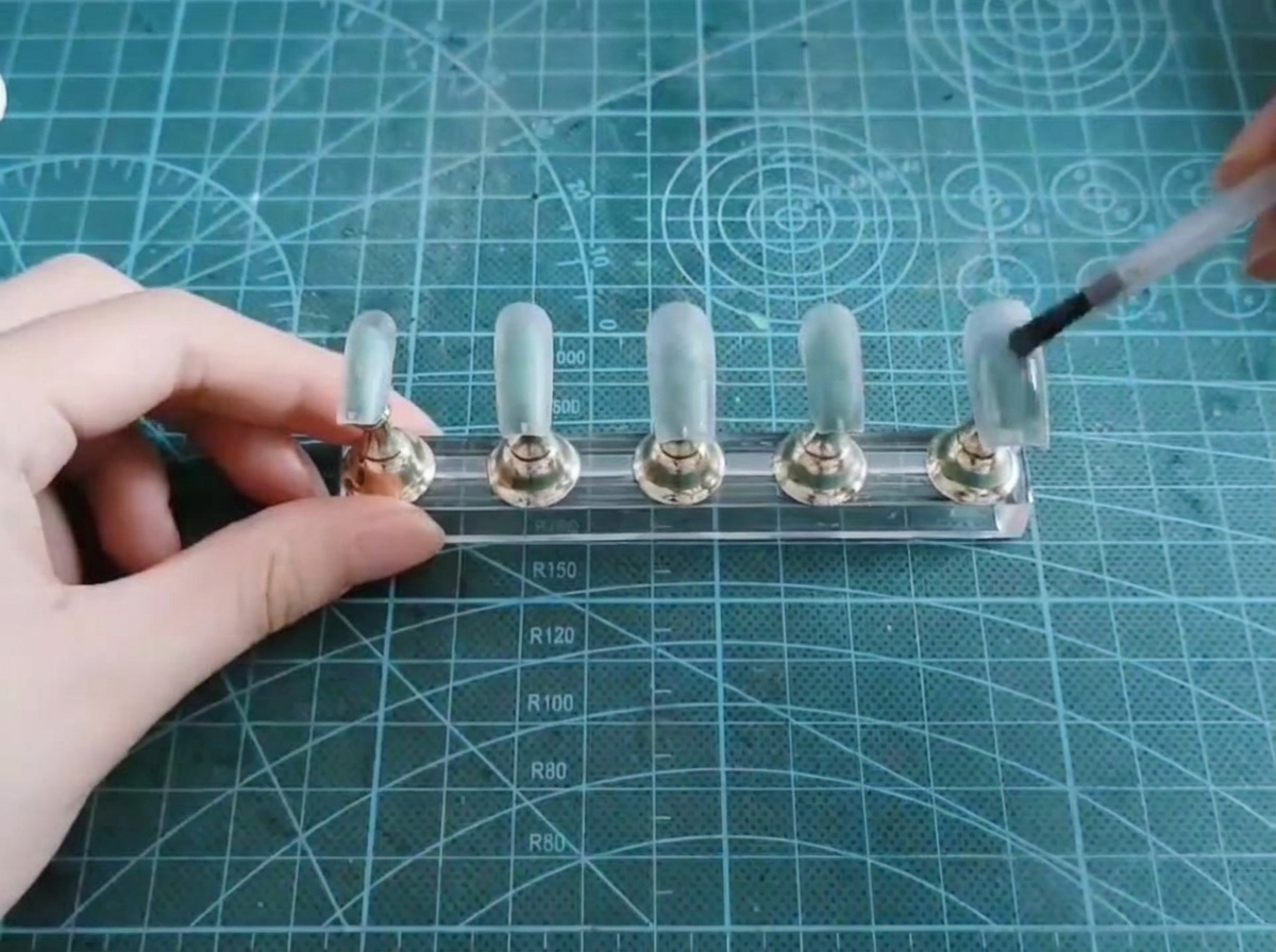 Ein Pinsel trägt eine Schicht transparentes Cat-Eye-Gel auf eine künstliche Nagelspitze auf.