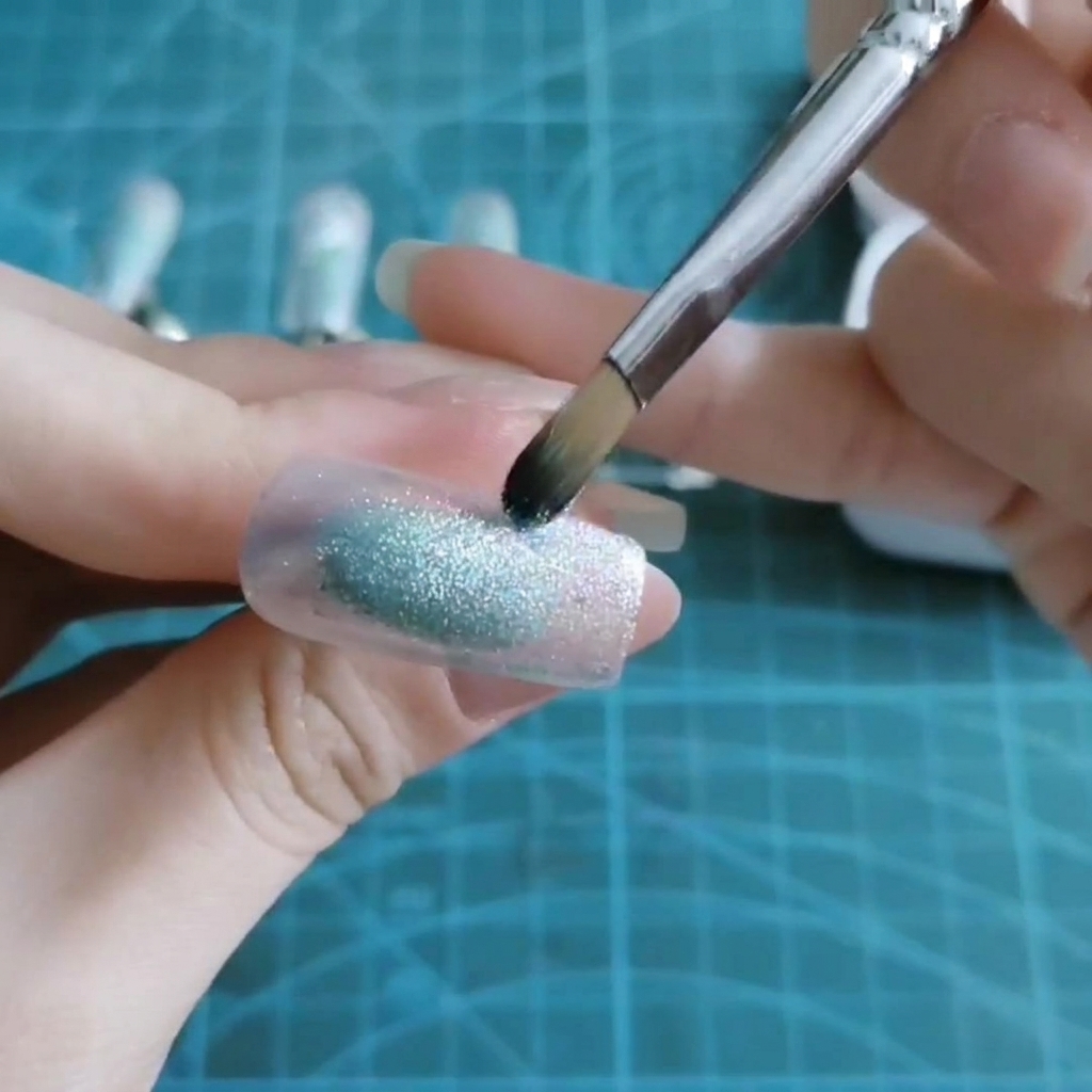 Ein Pinsel tupft transparentes blaues Gel auf die Mitte einer Nagelspitze.