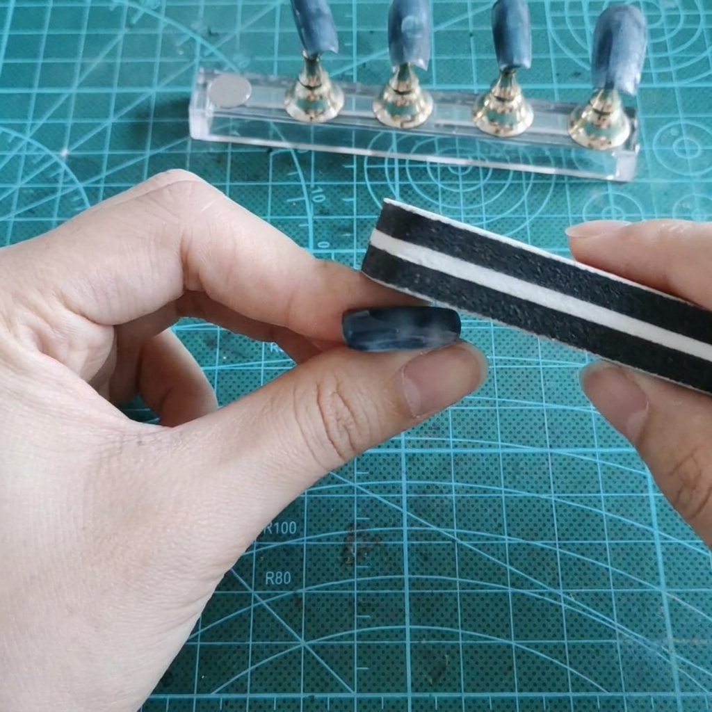 Ein Block-Schwamm-Buffer schleift die glänzende Geloberfläche eines künstlichen Nagels ab, der zwischen den Fingern gehalten wird.
