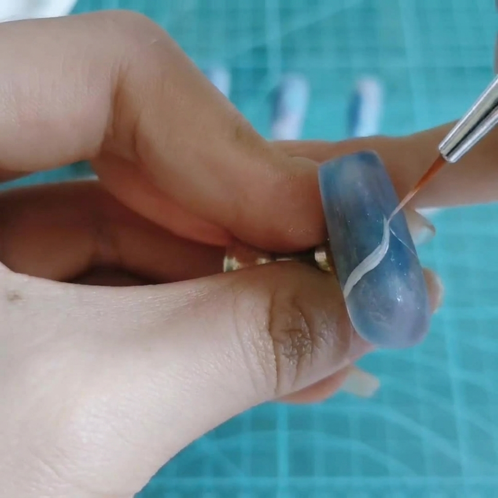 Ein feiner Nail-Art-Pinsel zieht eine dünne weiße, wellenförmige Linie über einen blau-lila marmorierten Nagel.