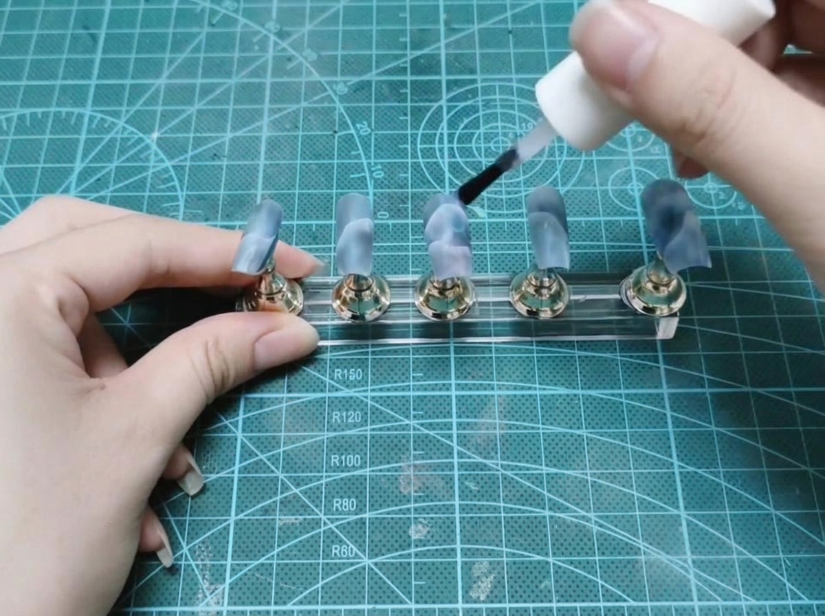 Ein Flaschenpinsel trägt klaren Aufbau-Gel auf einen Satz bemalter künstlicher Nägel auf einem Display-Ständer auf.