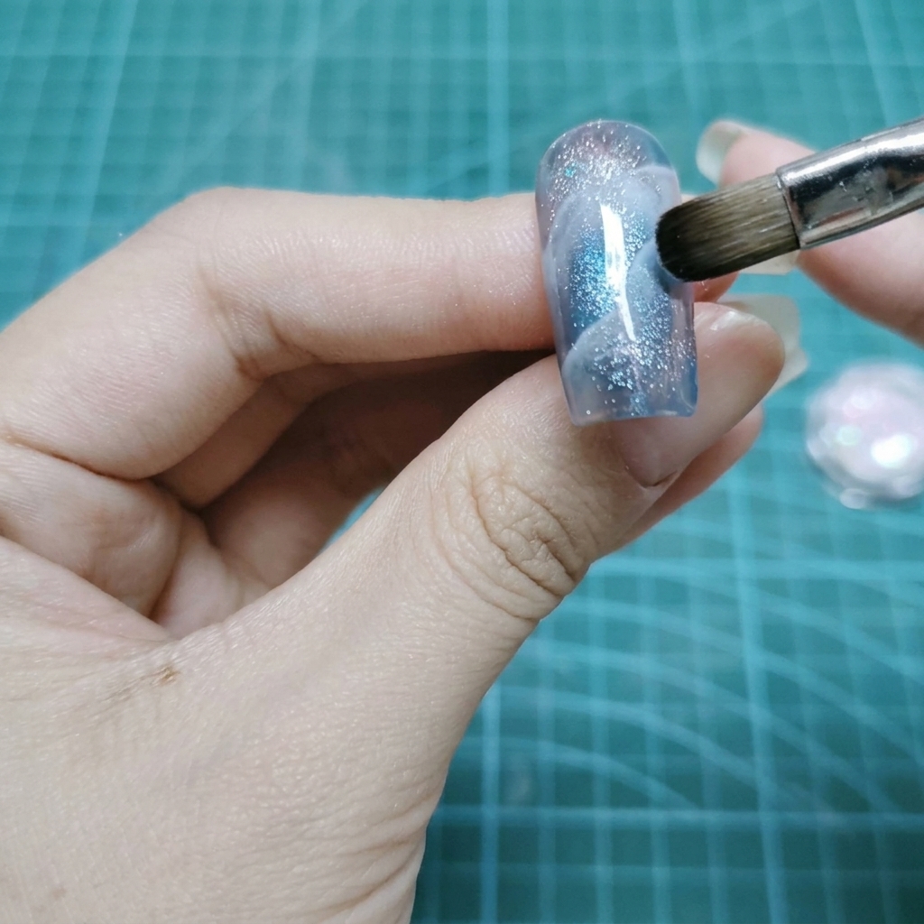 Ein flacher Make-up-Pinsel tupft holografische Glitzerpartikel auf eine feuchte Gel-Nageloberfläche.