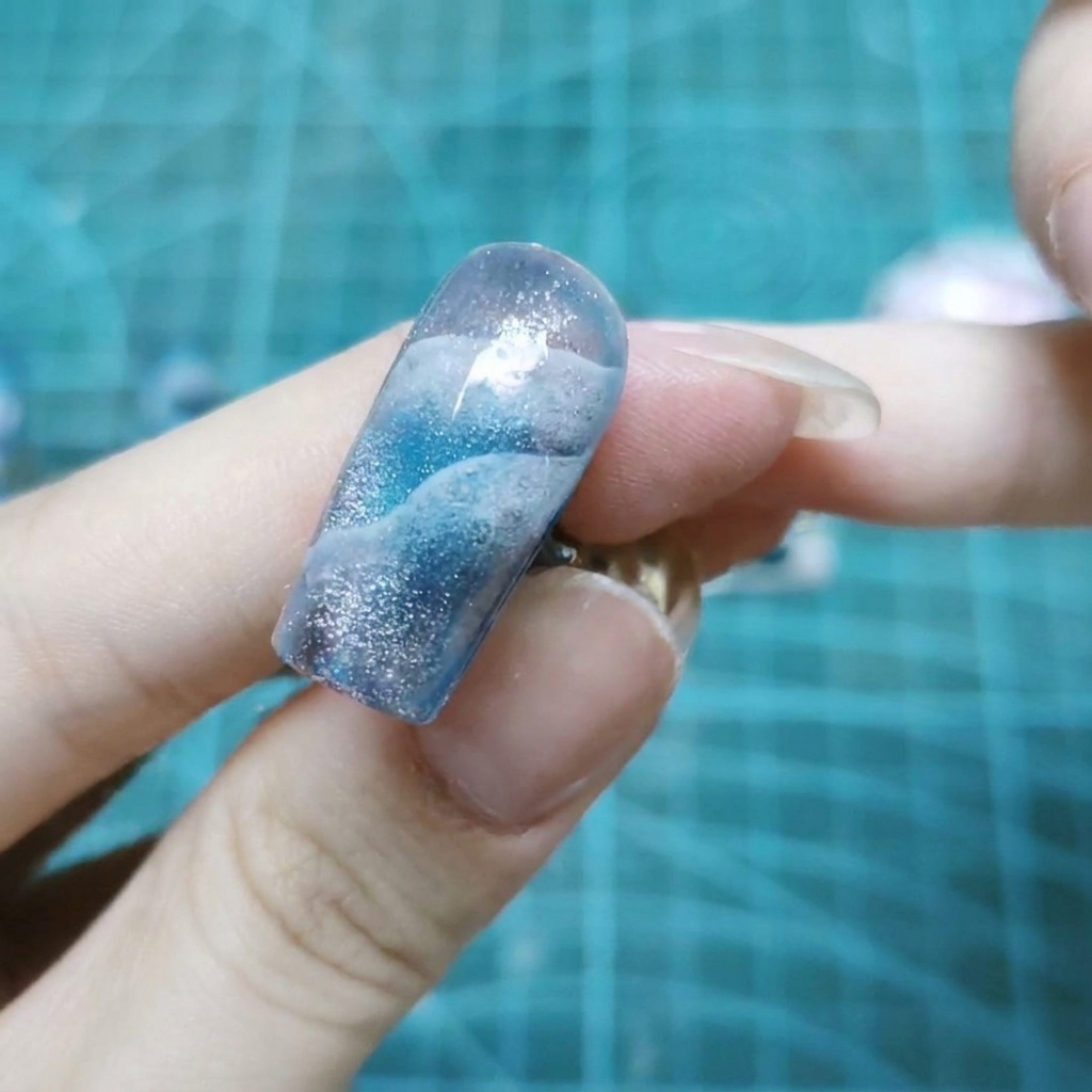 Eine Nahaufnahme eines einzelnen Nagels, der mit glitzernden holografischen Glitzerpartikeln bedeckt ist, die in klarem Gel eingebettet sind.