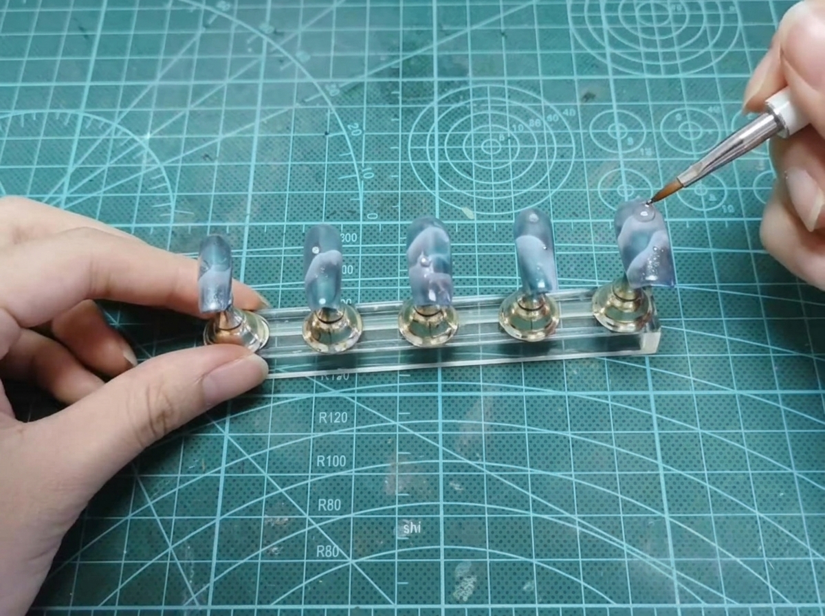 Ein Dotting-Tool platziert eine winzige silberne Perle in der Nähe des Nagelbettbereichs eines Nagels auf einem Ständer.