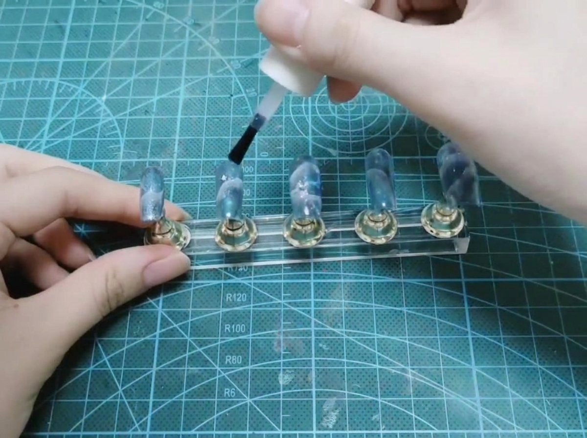 Eine Hand verwendet einen Pinsel aus einer weißen Flasche, um glänzenden Top Coat über eine Reihe fertiger Nägel auf einem Display-Ständer aufzutragen.