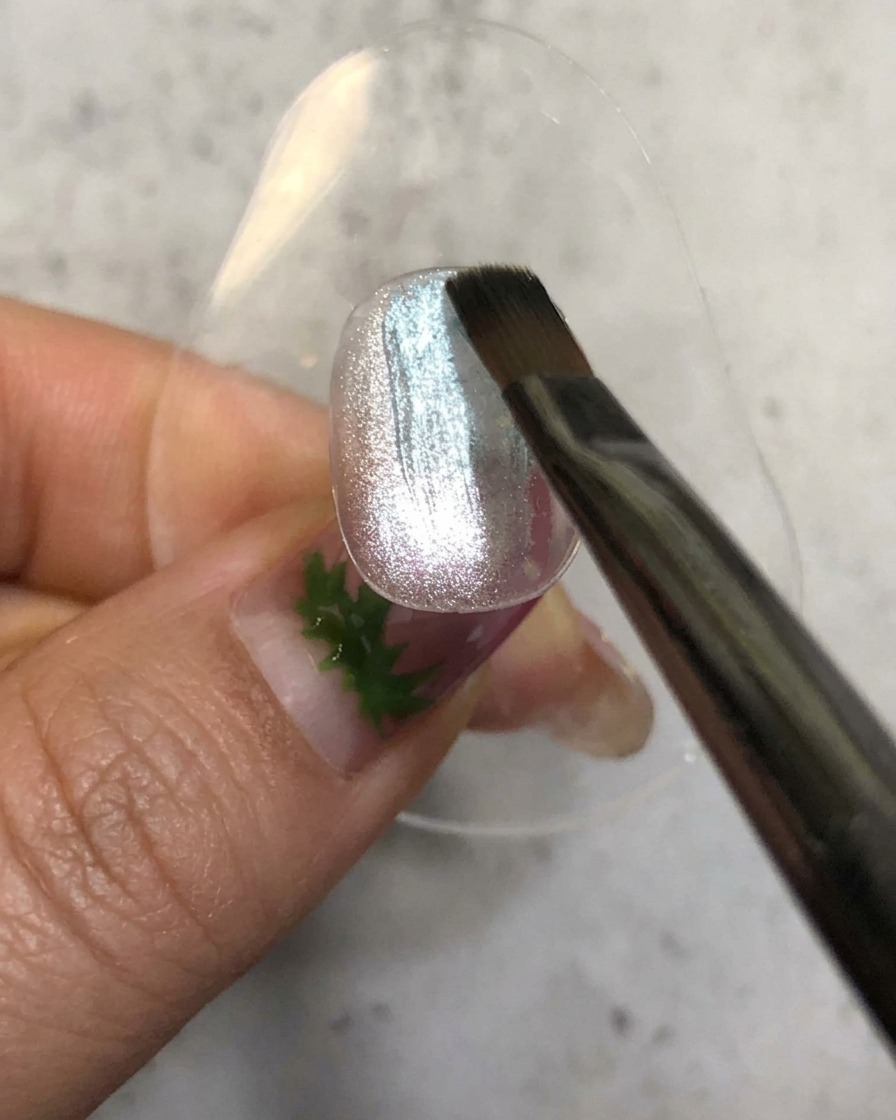 Ein Nagelpinsel trägt einen transparenten blauen Gellack über einer glitzernden silbernen Magnetbasis auf.