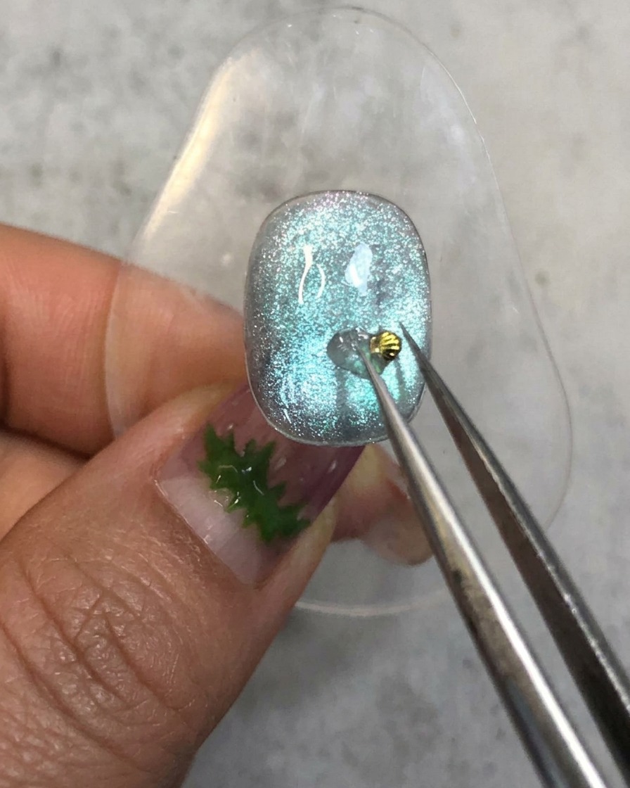 Eine Pinzette platziert ein winziges goldenes Muschel-Charm auf dem Aufbau-Gel-Punkt auf einem blauen Nagel.