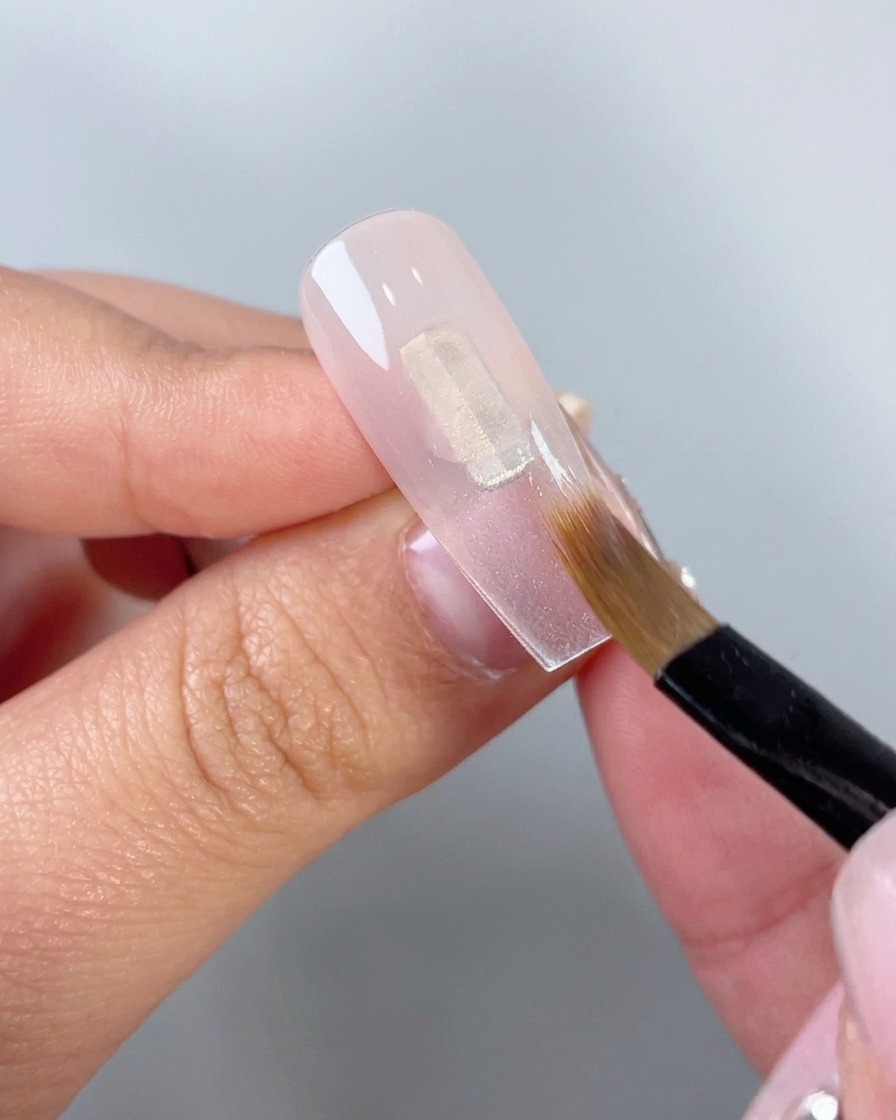 Eine Nahaufnahme eines flachen Make-up-Pinsels, der durchscheinendes rosa Gel nach unten auf einer klaren künstlichen Nagelspitze verblendet.