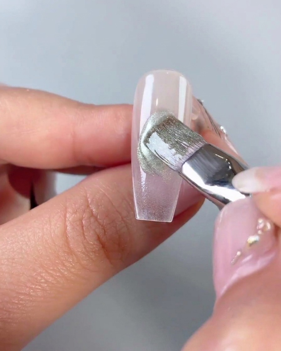 Ein flacher Pinsel, der grünes Cat-Eye-Gel glättet und verblendet, das diagonal über die Mitte einer klaren Nagelspitze aufgetragen wurde.