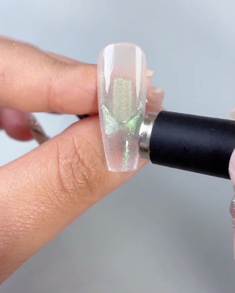 Ein schwarzer Magnetstab, der nah an die Seite einer klaren Nagelspitze gehalten wird und aktivierte grüne Cat-Eye-Schmetterlingsflügelmuster zeigt.