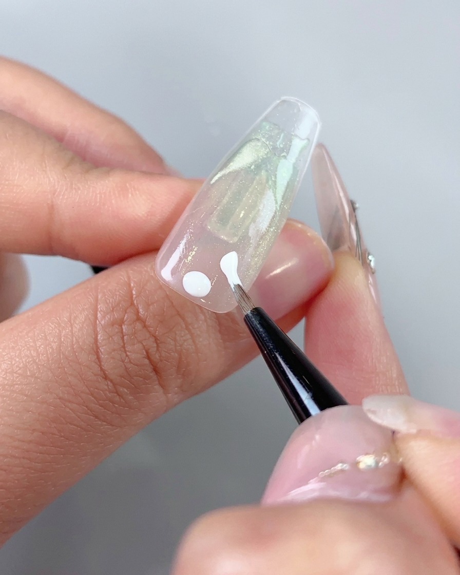 Ein feiner Kunstpinsel trägt weiße Gelpunkte und erste Striche nahe dem Nagelbettbereich eines Nagels auf.