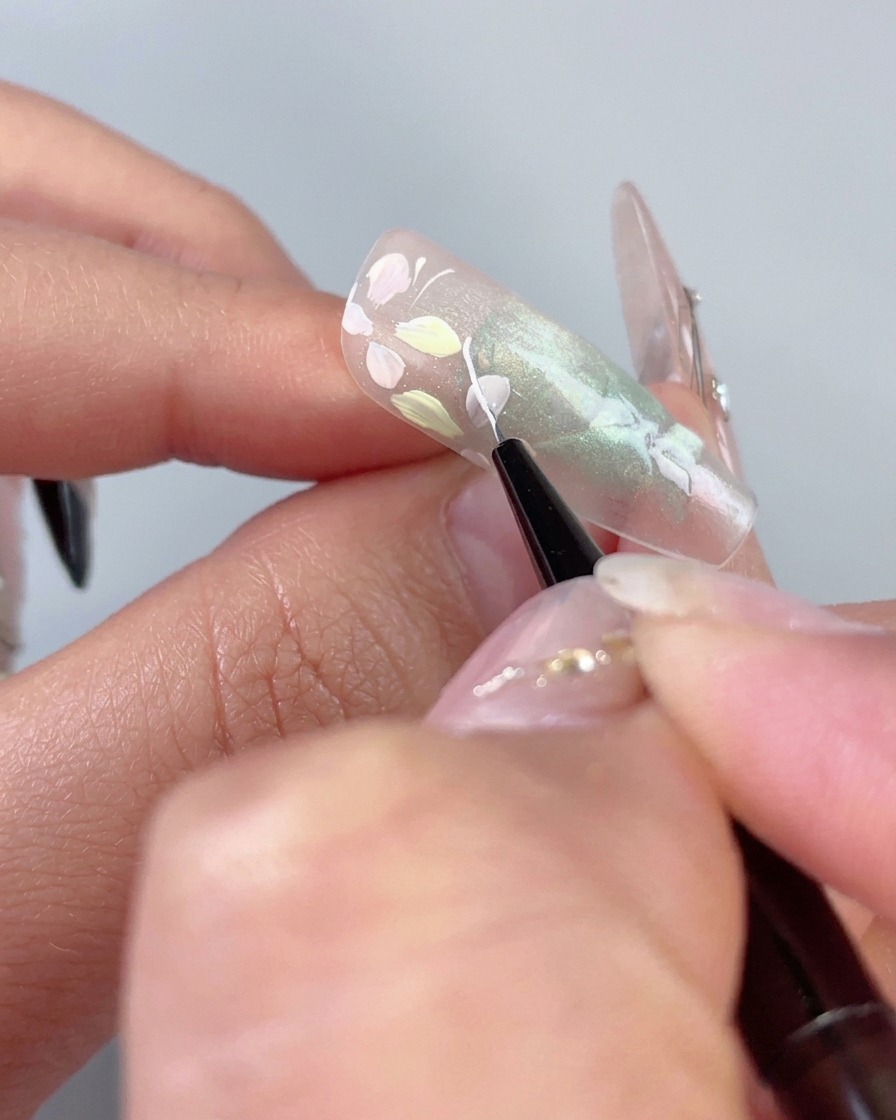 Ein feiner Liner-Pinsel zeichnet eine zarte weiße geschwungene Linie neben Pastellblumen auf einem durchsichtigen grünen Cat-Eye-Nagel.