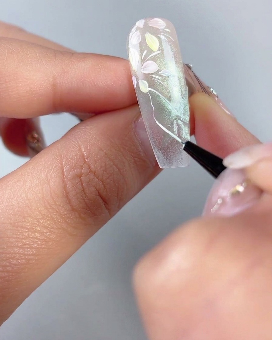 Ein feiner Liner-Pinsel zeichnet dünne weiße Umrisse und Flügelvenen über eine durchsichtige grüne Cat-Eye-Schmetterlingsform auf einem Nagel nach.
