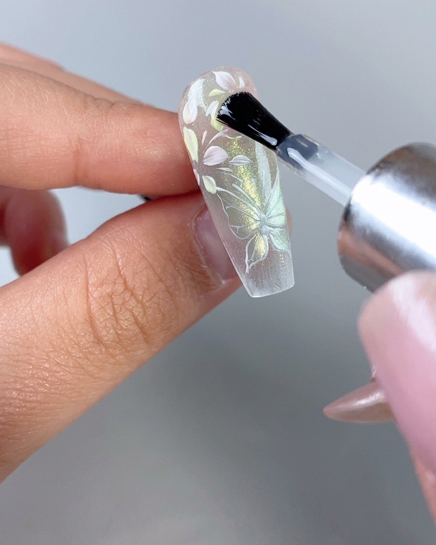 Ein Nagellackpinsel trägt einen glänzenden klaren Überzug über ein vollständig dekoriertes transparentes Blumen- und Schmetterlingsnagel auf.