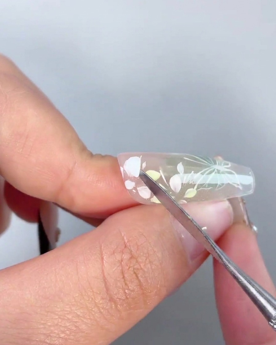 Ein Metallwerkzeug trägt einen Tropfen klaren Haftgels auf die Mitte eines gemalten Blumendesigns auf einem Nagel auf.