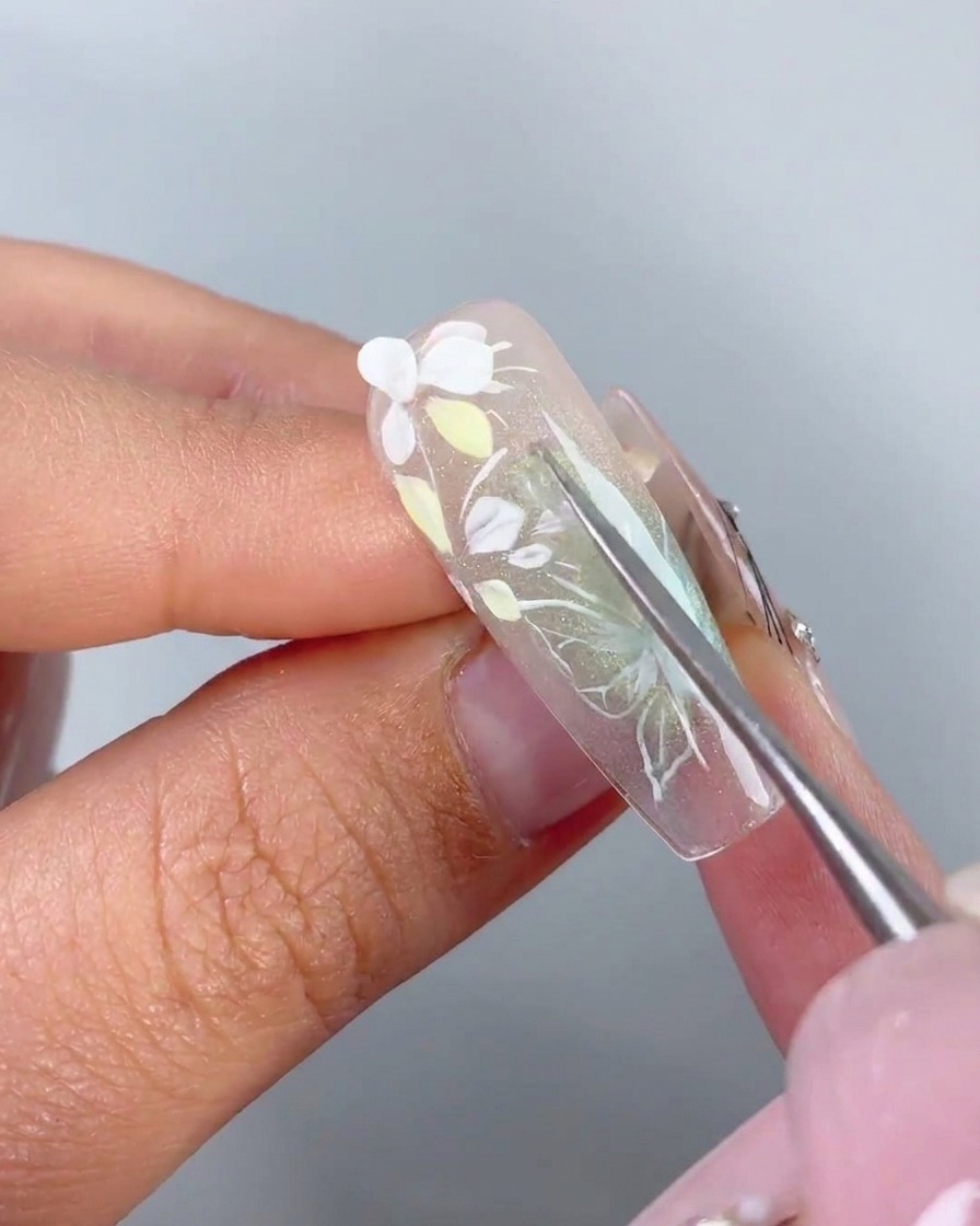 Ein Metallwerkzeug platziert ein vorgeformtes weißes 3D-Blütenblatt auf dem Haftgel eines Nagels.