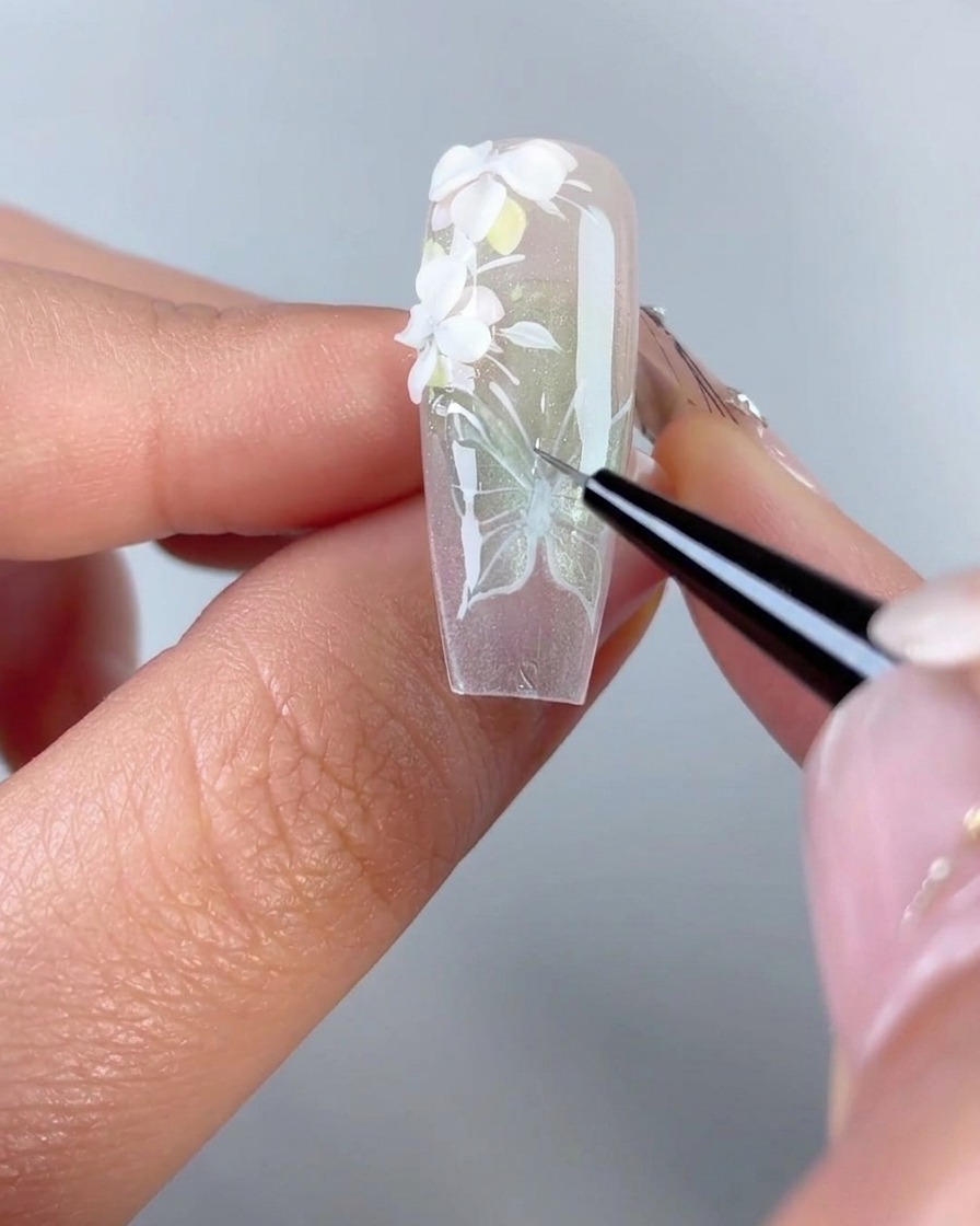 Eine Nahaufnahme eines feinen Liner-Pinsels zieht klares 3D-Modelliergel über die weiß gemalten Linien eines Schmetterlingsflügeldesigns auf einer Gelnagelspitze nach.