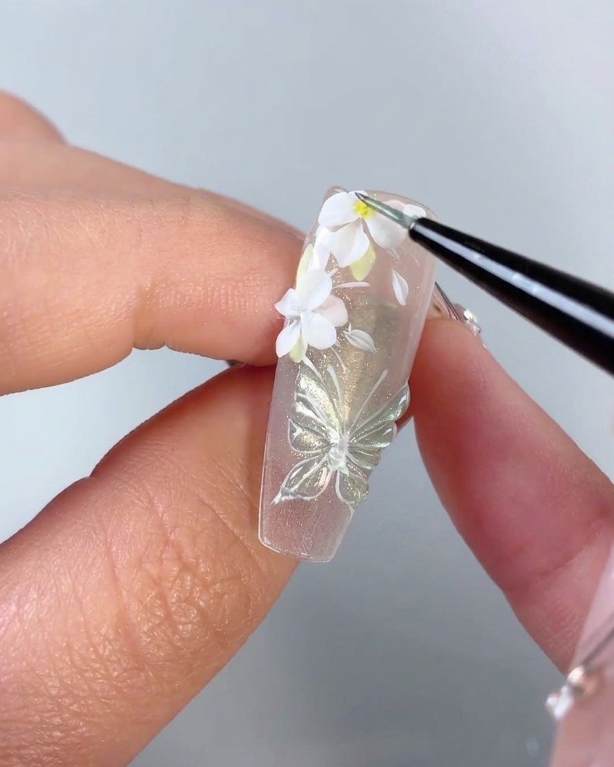 Ein feiner Pinsel fügt einen winzigen gelben Punkt in die Mitte einer weißen 3D-Blume auf einem Nagel hinzu.