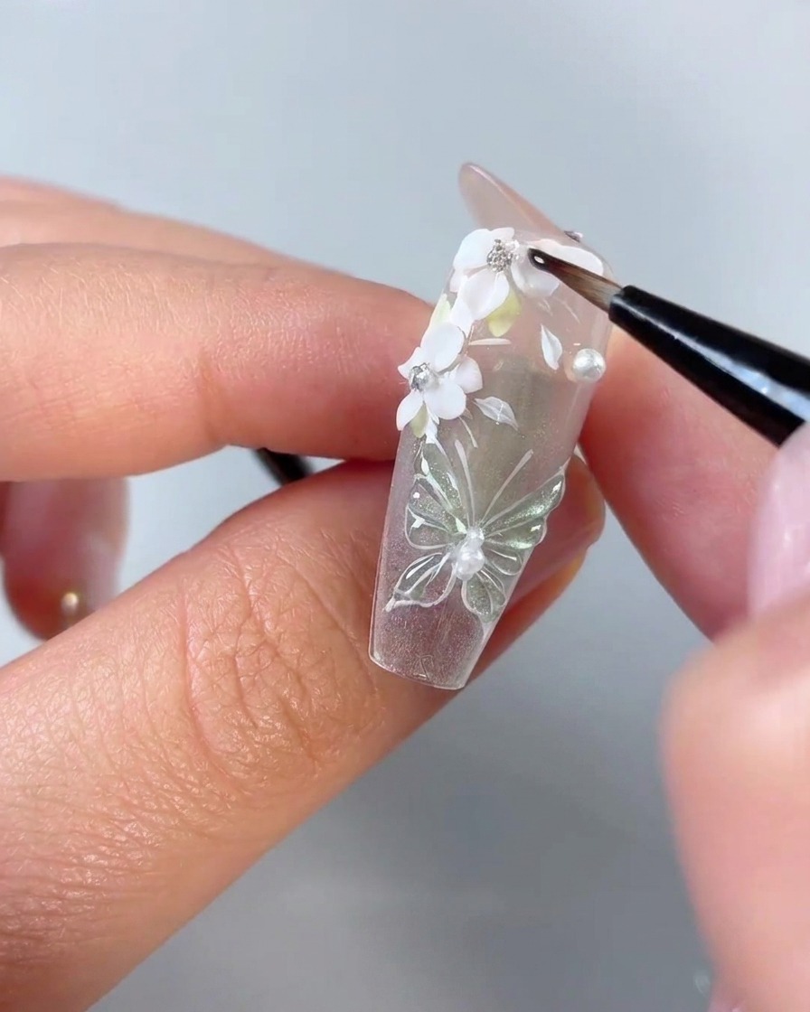 Ein feiner Detailnagelpinsel trägt klares Gel um die Kanten von weißen 3D-Blütenblättern und Strasssteinen auf einer klaren Nagelspitze auf.