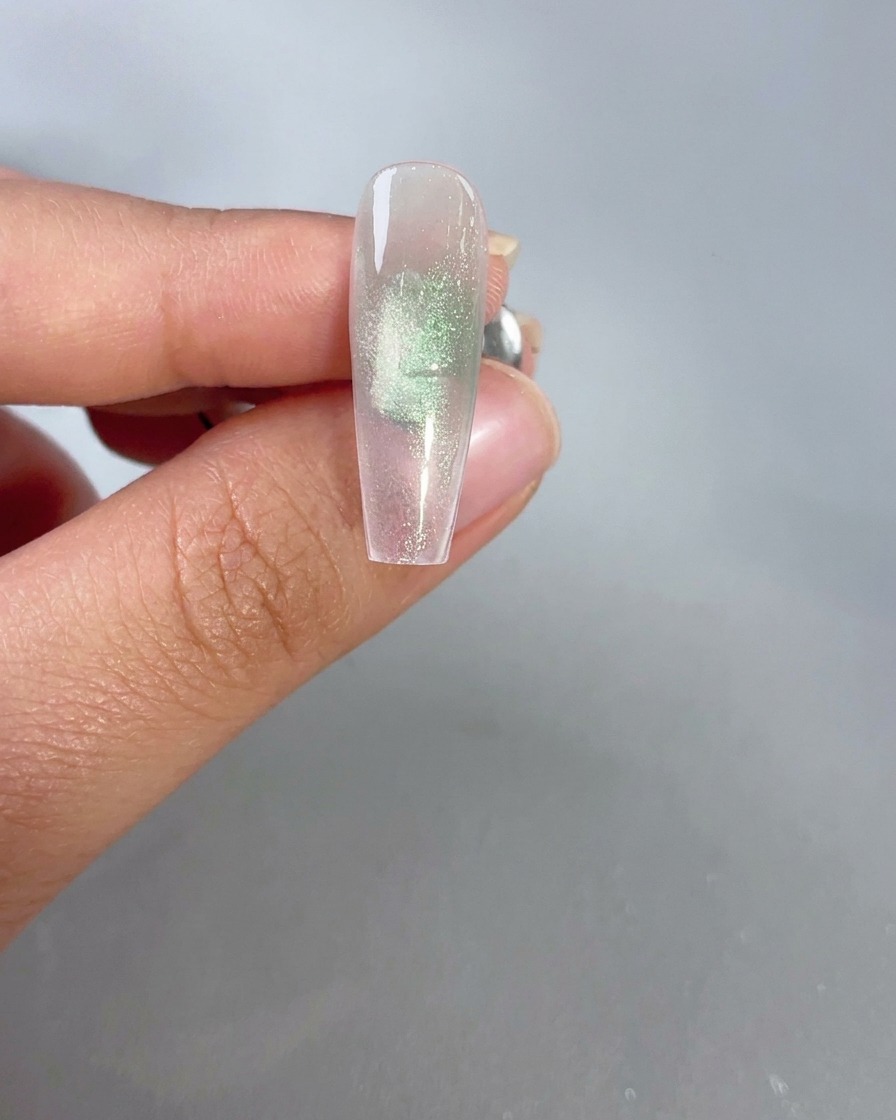 Ein Magnet wird nahe an einen durchsichtigen grünen Cat-Eye-Gelnagel gehalten, um den Shimmereffekt zu aktivieren.