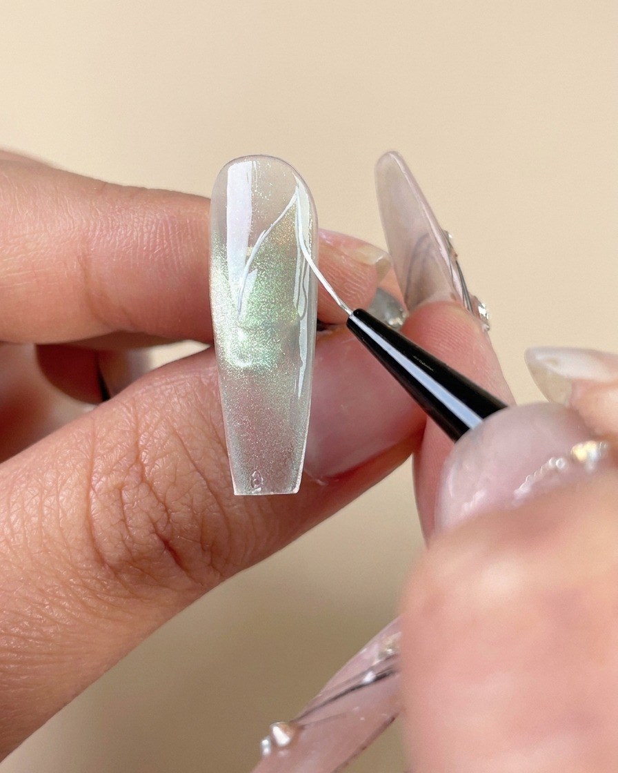 Ein feiner Liner-Pinsel zeichnet weiße Adernlinien auf einem grünen Cat-Eye-Gelnagel.