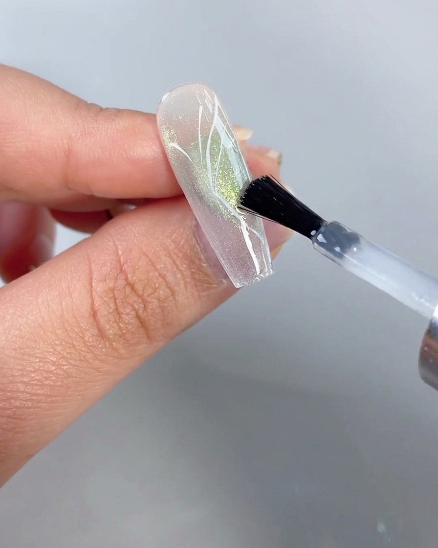 Ein Pinsel trägt klaren Überzug über einen Nagel mit einem gemalten Halbschmetterlingsdesign vor einem durchsichtigen grünen Cat-Eye-Hintergrund auf.