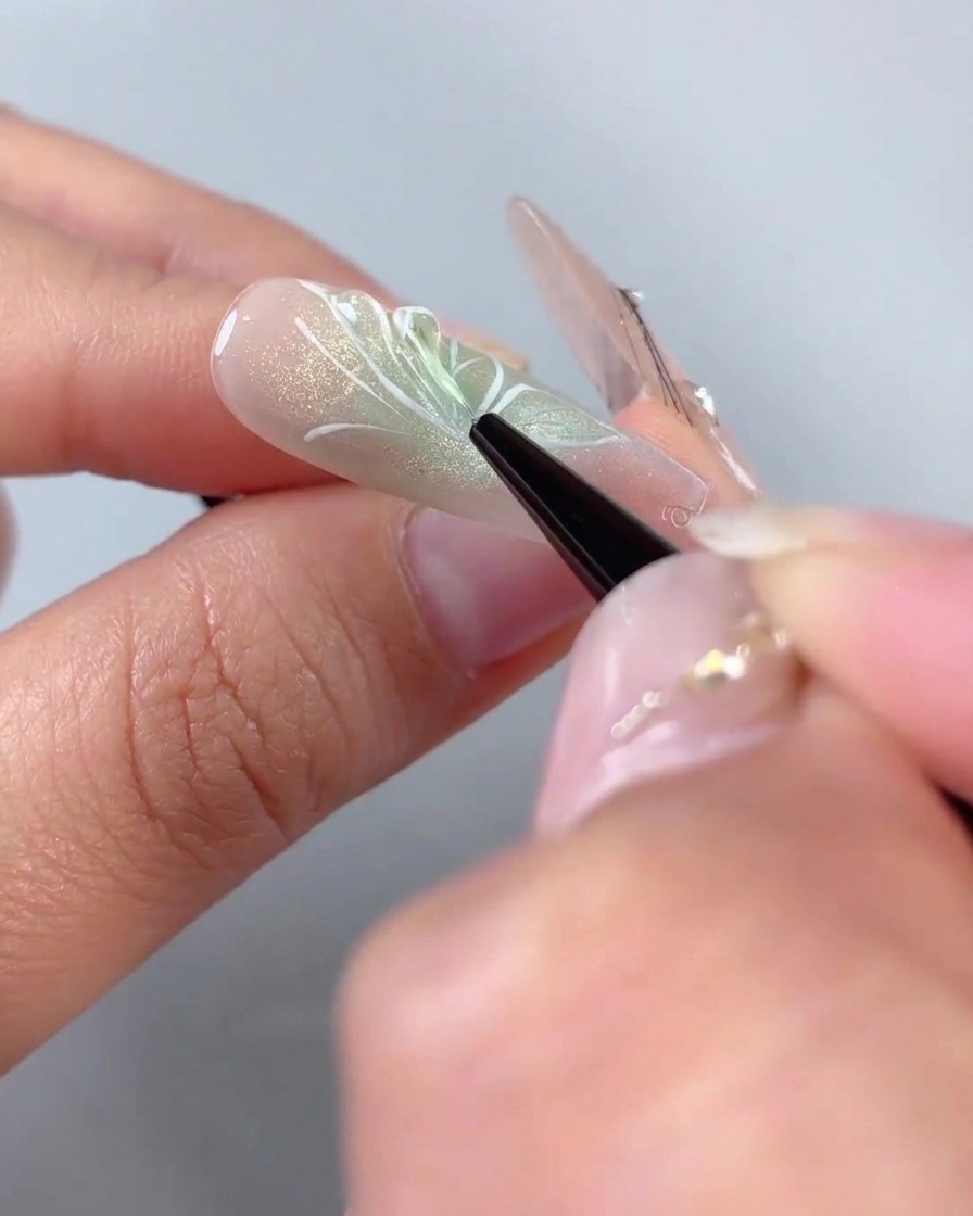 Ein feiner Liner-Pinsel trägt klares 3D-Modelliergel über die weiß gemalten Flügelumrisse eines Halbschmetterlings-Nageldesigns auf.