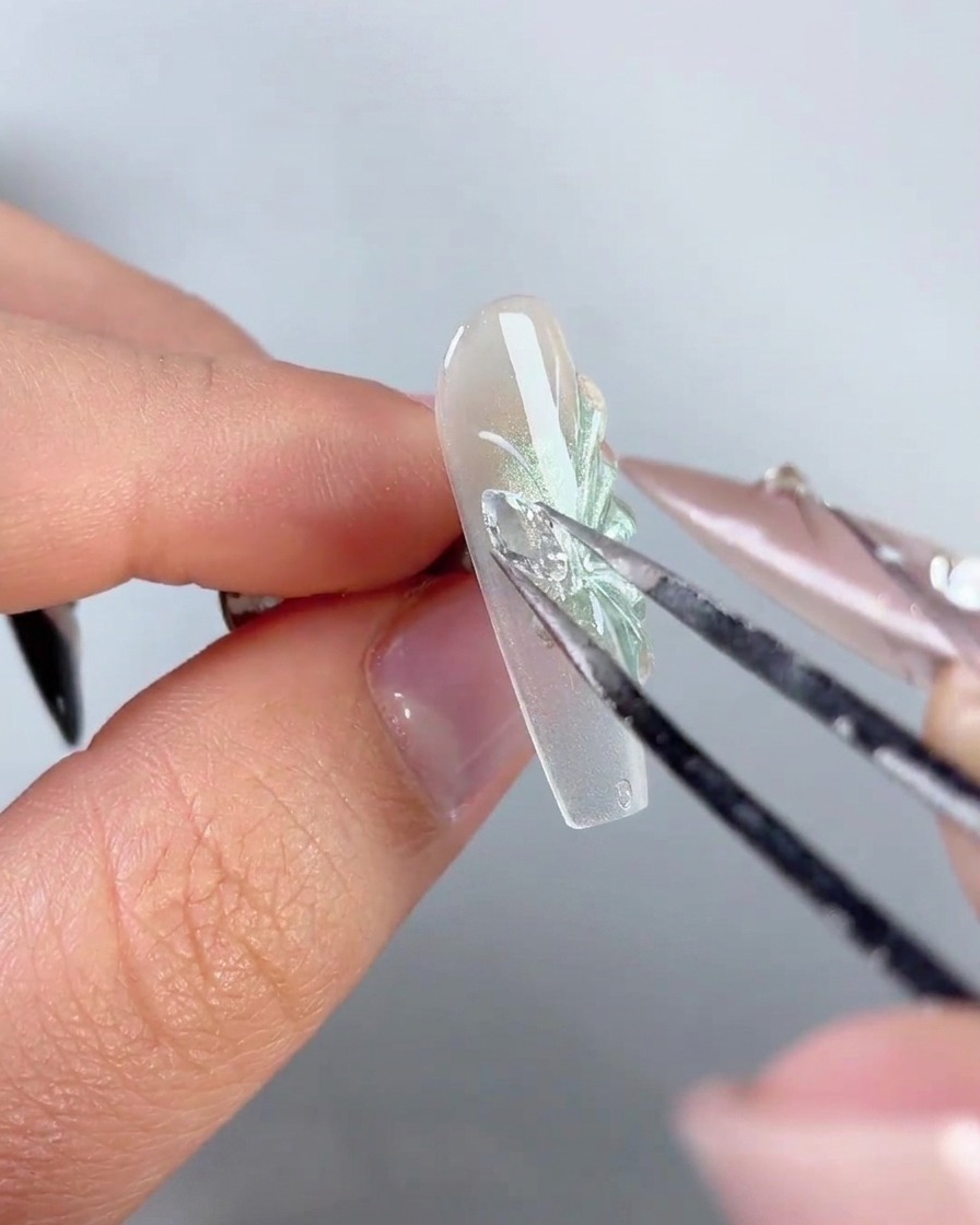 Eine Metallpinzette platziert einen glitzernden Strass-Schmetterlingsflügel-Charm auf die Seite eines dekorierten Nagels.